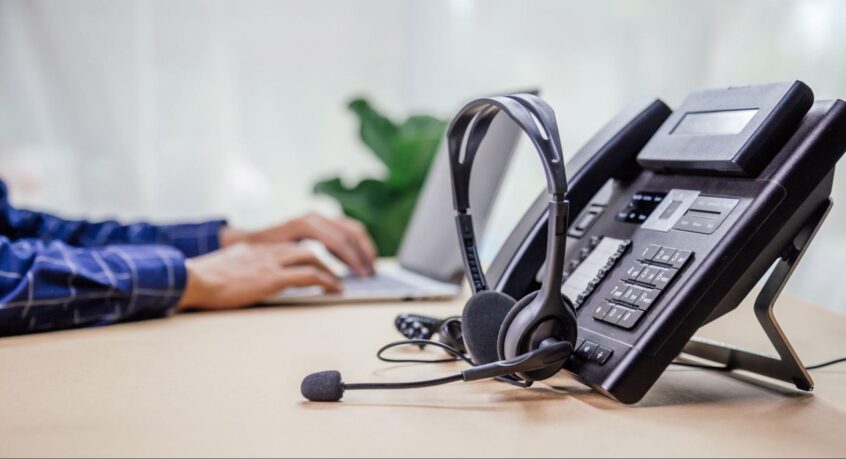 Office desk with a business phone, headset, and employee working on a laptop in the background.