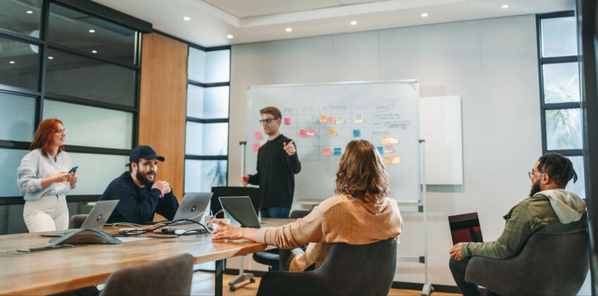 Team meeting in a modern conference room with a presenter standing by a whiteboard and colleagues gathered around a table with laptops.